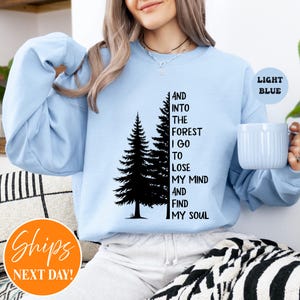 May include: A light blue crewneck sweatshirt featuring a black graphic of two pine trees and the text, "AND INTO THE FOREST I GO TO LOSE MY MIND AND FIND MY SOUL." The garment has long sleeves and a ribbed neckline, providing a simple, casual appearance.