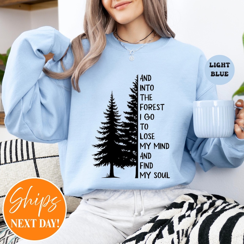 May include: A light blue crewneck sweatshirt featuring a black graphic of two pine trees and the text, "AND INTO THE FOREST I GO TO LOSE MY MIND AND FIND MY SOUL." The garment has long sleeves and a ribbed neckline, providing a simple, casual appearance.