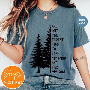 May include: A blue-gray short-sleeve t-shirt featuring a black graphic of two pine trees and the text: AND INTO THE FOREST I GO TO LOSE MY MIND AND FIND MY SOUL. The shirt has a crew neckline and is worn with blue denim jeans.