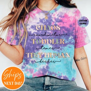 May include: A short-sleeve t-shirt with a pink, purple, and blue tie-dye pattern. The front features the text: CHAOS coordinator, TODDLER tamer, TINY HUMAN herder. A small circular badge in the top right corner reads COTTON CANDY.