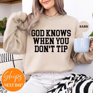 May include: A sand-colored crewneck sweatshirt featuring the black text "GOD KNOWS WHEN YOU DON'T TIP" printed on the front. The long-sleeved garment has a ribbed neckline, cuffs, and hem, and is worn with light-colored pants.