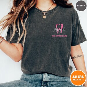 May include: Dark gray t-shirt with a pink tooth graphic and the name "Abigail" in cursive. Below the name is the text "YOUR CORPORATE NAME" in pink. The shirt is a crew neck style.