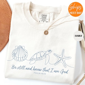 May include: Ivory-colored t-shirt with a blue seashell, turtle, and starfish design. The text "Be still and know that I am God, Psalm 46:10" is printed below the design. A gold necklace is draped on the shirt.