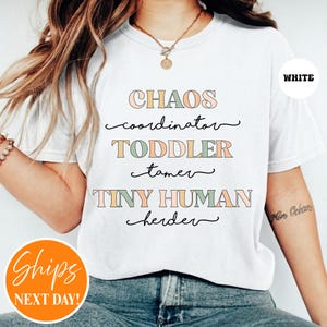May include: A white t-shirt featuring the text CHAOS coordinator, TODDLER tamer, and TINY HUMAN herder in a mix of block and cursive fonts. The block letters are colored in muted shades of pink, green, and orange, while the cursive text is black.
