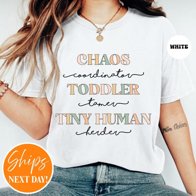 May include: A white t-shirt featuring the text CHAOS coordinator, TODDLER tamer, and TINY HUMAN herder in a mix of block and cursive fonts. The block letters are colored in muted shades of pink, green, and orange, while the cursive text is black.