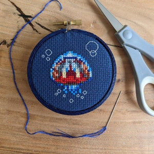 May include: A blue and red cross-stitch embroidery of a pixelated character with a large mouth, surrounded by white dots on a dark blue background.