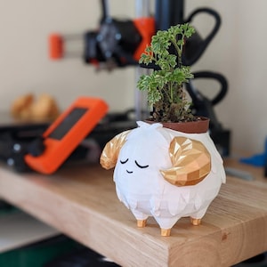 May include: A white 3D printed planter shaped like a fluffy sheep with gold horns. The planter is holding a small green plant.