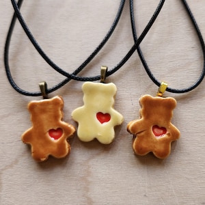 May include: Three ceramic teddy bear pendants with red heart accents on black cords. The pendants are in shades of brown and cream, with gold and bronze-colored clasps. The background is a light wood grain.