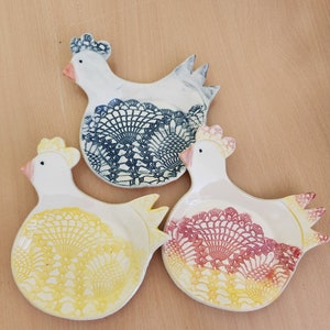 May include: Three ceramic chicken-shaped dishes with lace designs. The dishes are white with blue, yellow, and red lace patterns.