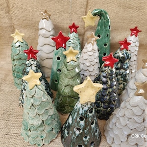 May include: A collection of ceramic Christmas trees in various shades of green, white, and blue. Each tree is topped with a star, either red or yellow. The trees have textured surfaces and are arranged on a neutral background.