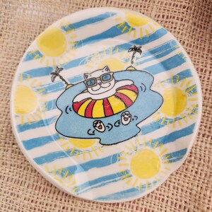 May include: A round, white ceramic plate with a whimsical design. The plate features a cartoon cat wearing sunglasses and floating in a pool on an inner tube. The background has blue stripes, yellow suns, and palm trees. The plate is approximately 6 inches in diameter.