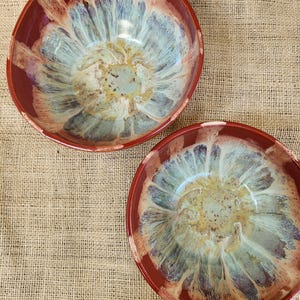 May include: Two handmade ceramic bowls with a floral design. The bowls have a deep red rim and a center with shades of blue, green, and yellow. The bowls are on a tan burlap surface. The text "CRK Ceramics" is in the bottom right corner.