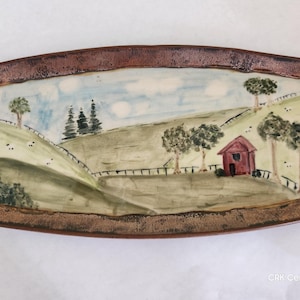 May include: A rectangular ceramic tray with a dark brown textured rim and a hand-painted landscape scene featuring a red barn, green rolling hills, trees, and a fence under a blue sky with clouds. The text CRK Ceramics is in the bottom corner.