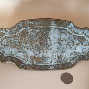 May include: A light blue ceramic serving dish with a raised design of sea creatures, including sea turtles, starfish, and shells. The dish has a scalloped edge and a brown glaze.