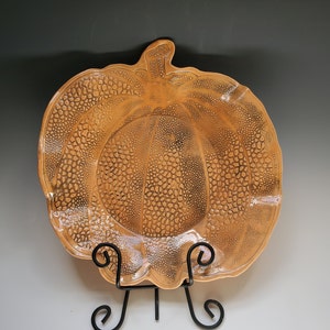 May include: A ceramic pumpkin-shaped serving dish with a textured surface. The dish is a light orange color and has a scalloped edge. It is displayed on a black metal stand.