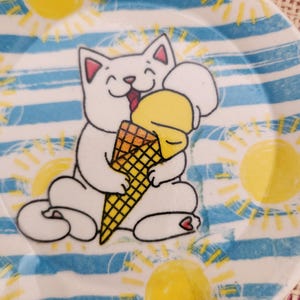 May include: A white ceramic plate with a cartoon cat hugging a yellow ice cream cone. The plate has blue and white stripes, with yellow sun illustrations. The cat has pink ears and a smiling face. A fun, summer-themed plate.