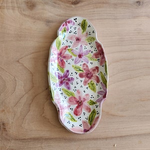 May include: A white ceramic serving dish with a floral design. The dish is oval shaped and features pink and purple flowers with green leaves and black dots.
