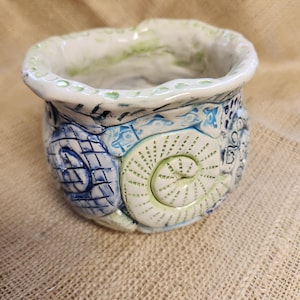 May include: A white ceramic planter with a green, blue, and white seashell design. The planter has a slightly wavy rim and is decorated with raised seashells.