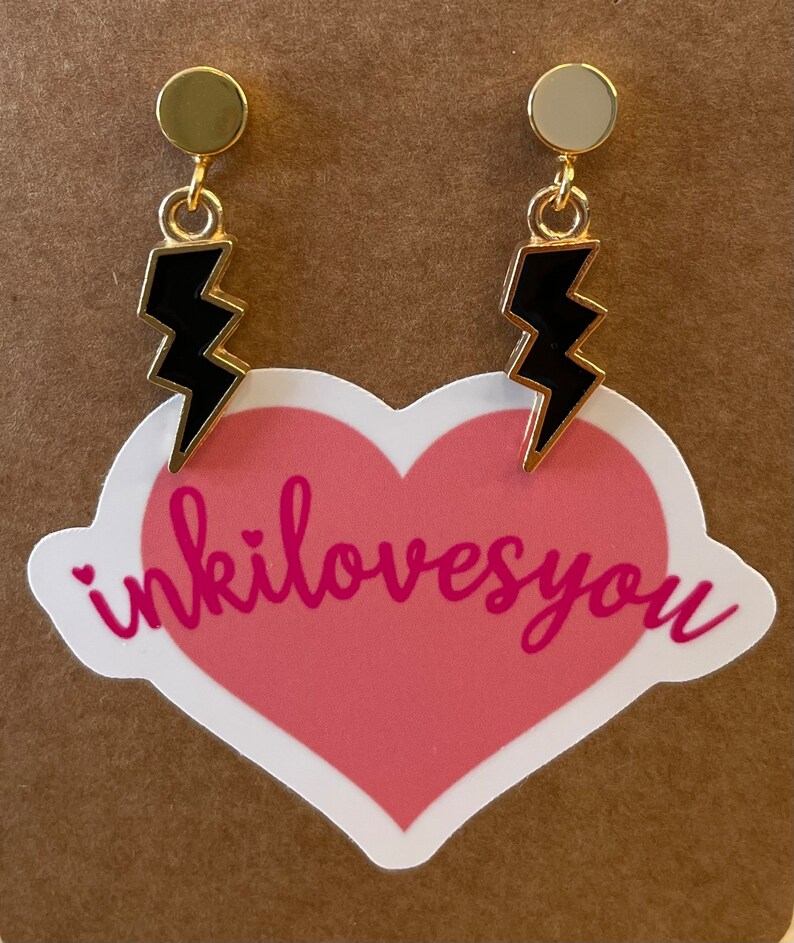 May include: A pair of gold earrings with black lightning bolt charms. The earrings have a simple, minimalist design and are perfect for adding a touch of edgy style to any outfit.