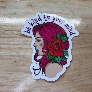 May include: A sticker with a pink-haired woman with a red rose and green leaves behind her. The text "be kind to your mind" is written in a purple font around the woman.