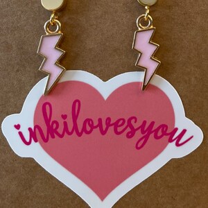 Pode incluir: Um par de brincos de ouro com pingentes de raio rosa. Os brincos est&atilde;o em um adesivo em forma de cora&ccedil;&atilde;o rosa com o texto "inkilovesyou".