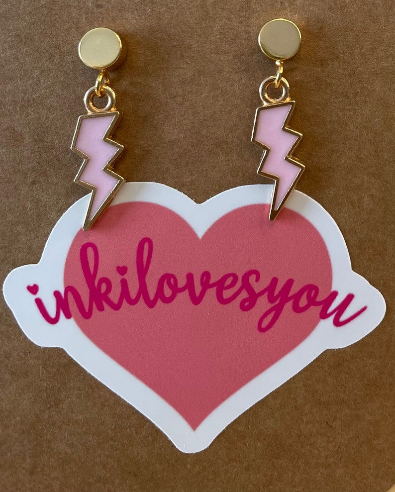 Pode incluir: Um par de brincos de ouro com pingentes de raio rosa. Os brincos est&atilde;o em um adesivo em forma de cora&ccedil;&atilde;o rosa com o texto "inkilovesyou".