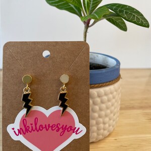 May include: A pair of gold stud earrings with black lightning bolt charms. The earrings are displayed on a brown card with a pink heart sticker that says "inkilovesyou".