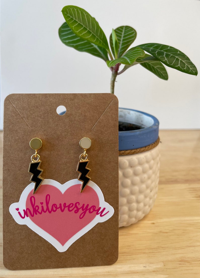 May include: A pair of gold stud earrings with black lightning bolt charms. The earrings are displayed on a brown card with a pink heart sticker that says "inkilovesyou".
