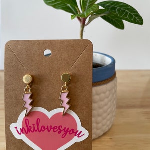 Pode incluir: Um par de brincos de raio dourado com esmalte rosa. Os brincos est&atilde;o em um cart&atilde;o marrom com um adesivo de cora&ccedil;&atilde;o rosa que diz "inkilovesyou".