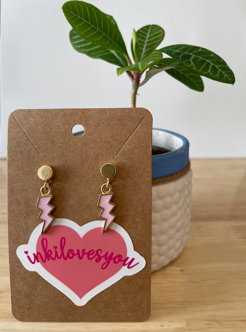 Pode incluir: Um par de brincos de raio dourado com esmalte rosa. Os brincos est&atilde;o em um cart&atilde;o marrom com um adesivo de cora&ccedil;&atilde;o rosa que diz "inkilovesyou".