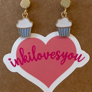 May include: A pair of gold cupcake earrings with white frosting and blue icing. The earrings are on a brown background with a pink heart sticker that says "inkilovesyou".