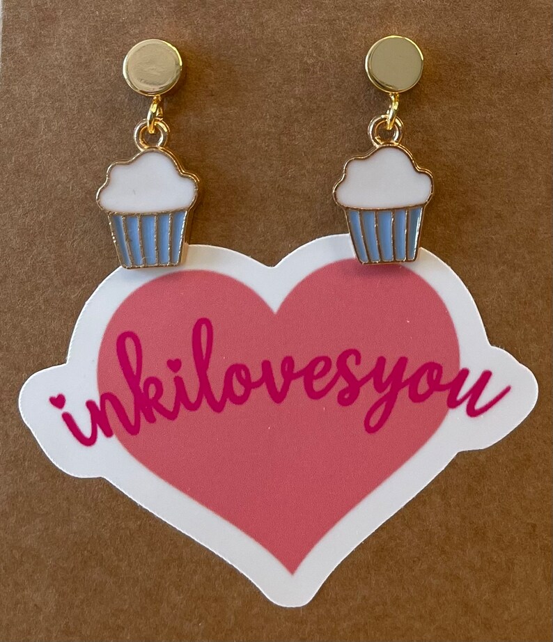 May include: A pair of gold cupcake earrings with white frosting and blue icing. The earrings are on a brown background with a pink heart sticker that says "inkilovesyou".