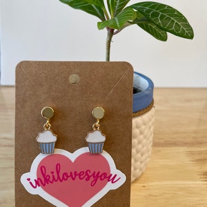 May include: A pair of gold cupcake earrings with white frosting and blue icing. The earrings are displayed on a brown card with a pink heart sticker that says "inkilovesyou".