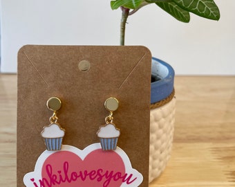 Pode incluir: Um par de brincos de cupcake dourados com cobertura branca e cobertura azul. Os brincos são exibidos em um cartão marrom com um adesivo de coração rosa que diz "inkilovesyou".