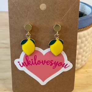 May include: A pair of gold lemon earrings with green leaves. The earrings are on a brown card with a pink heart sticker that says "inkilovesyou".