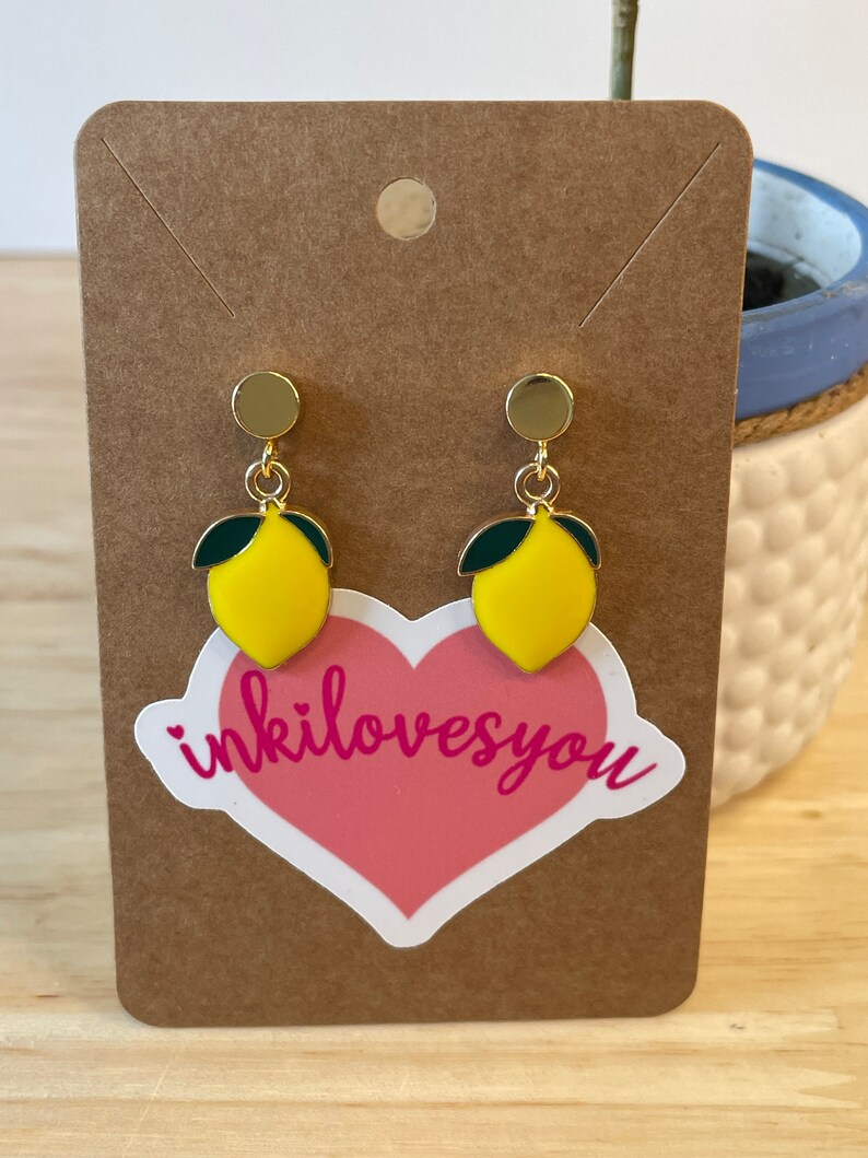 May include: A pair of gold lemon earrings with green leaves. The earrings are on a brown card with a pink heart sticker that says "inkilovesyou".