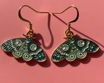 Pode incluir: Um par de brincos de metal dourado com um design de traça. As asas da traça são verde-azuis e brancas com um padrão de lua crescente e estrelas. Num fundo rosa.