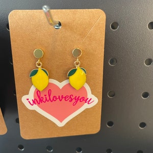 May include: A pair of gold lemon earrings with green leaves. The earrings are displayed on a brown card with a pink heart sticker that says "inkilovesyou".