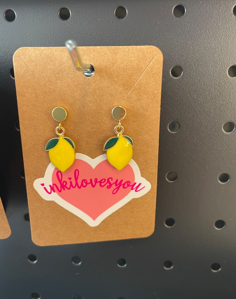 May include: A pair of gold lemon earrings with green leaves. The earrings are displayed on a brown card with a pink heart sticker that says "inkilovesyou".