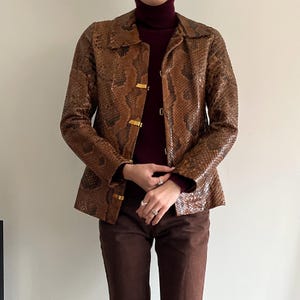 May include: Brown snakeskin-patterned jacket with gold-colored clasps. The jacket has a collared neckline and long sleeves. The model is wearing a burgundy turtleneck and brown pants. The jacket is a vintage style.