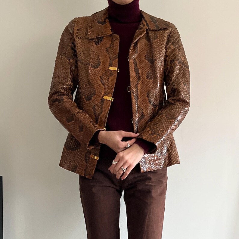 May include: Brown snakeskin-patterned jacket with gold-colored clasps. The jacket has a collared neckline and long sleeves. The model is wearing a burgundy turtleneck and brown pants. The jacket is a vintage style.
