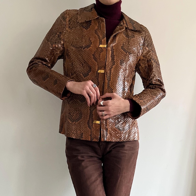 May include: Brown snakeskin-patterned jacket with a collared neckline and gold-colored button closures. The jacket is worn over a burgundy turtleneck and paired with brown pants. The jacket's texture and color create a vintage aesthetic.