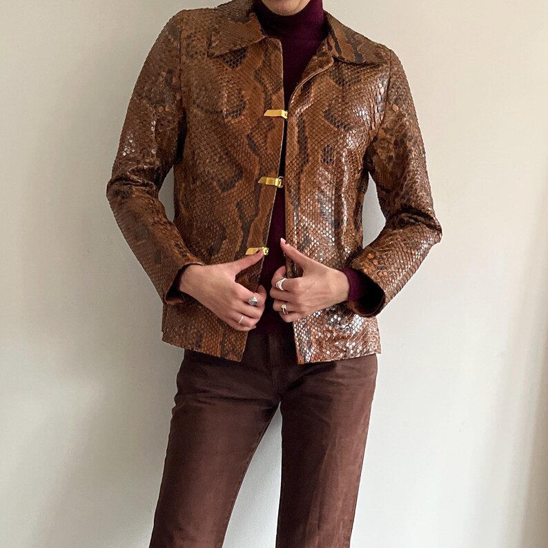 May include: Brown snakeskin-patterned jacket with gold-colored clasps. The jacket has a collared neckline and long sleeves. The model is wearing a burgundy turtleneck and brown pants.
