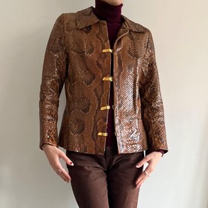 May include: Brown snakeskin-patterned jacket with gold-colored clasps. The jacket has a collared neckline and long sleeves. The garment is worn over a burgundy turtleneck and paired with brown pants. The jacket's texture and color create a vintage aesthetic.