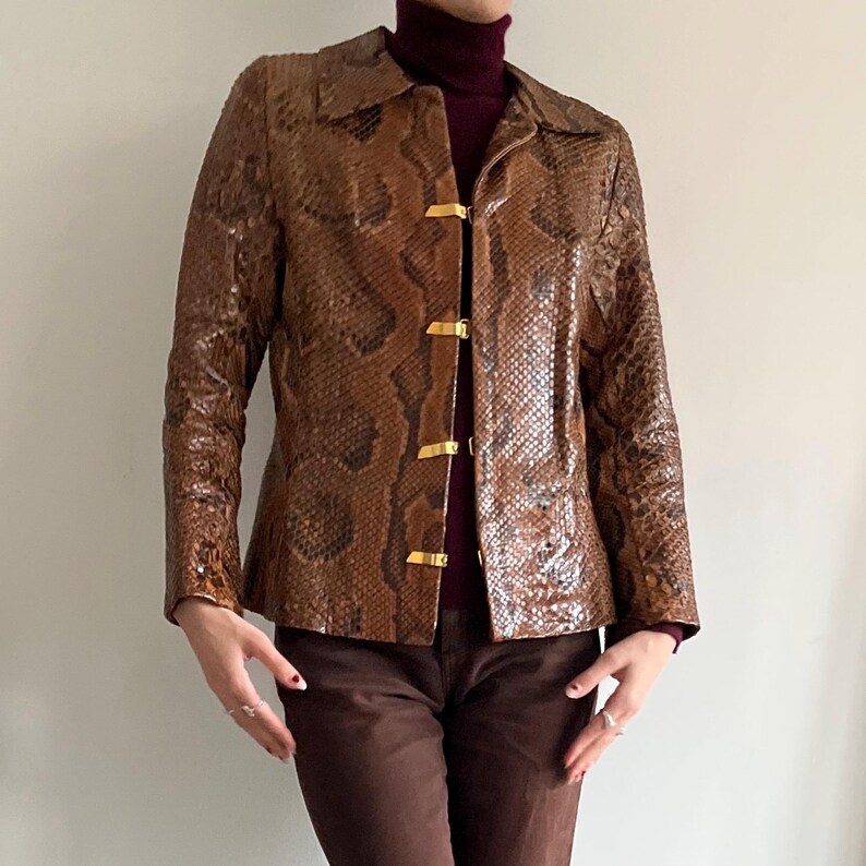 May include: Brown snakeskin-patterned jacket with gold-colored clasps. The jacket has a collared neckline and long sleeves. The garment is worn over a burgundy turtleneck and paired with brown pants. The jacket's texture and color create a vintage aesthetic.