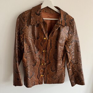 May include: Brown and black snakeskin-patterned jacket with a collared neckline and three gold-colored clasps. The jacket is hanging on a white hanger. The jacket has a brown interior lining.