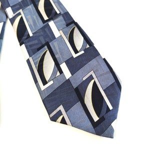 May include: A patterned necktie featuring a geometric design in shades of blue, gray, and white. The tie has a repeating pattern of squares and curved shapes, with a light-colored background.