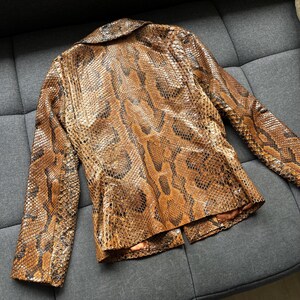 May include: A brown and tan snakeskin-patterned blazer with a notched collar and long sleeves. The jacket has a tailored fit and a two-button closure. The snakeskin pattern is detailed with dark brown and black accents.