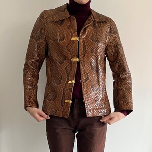 May include: Brown snakeskin-patterned jacket with gold-colored clasps. The jacket has long sleeves and a collared neckline. The jacket is worn over a burgundy turtleneck. The jacket is a vintage style.