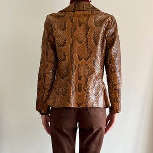 May include: A brown snakeskin-patterned blazer with a notched collar and long sleeves. The jacket has a fitted silhouette and is paired with brown pants. The snakeskin pattern features shades of brown and black.